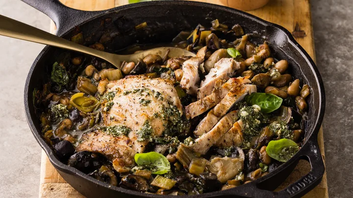 Skillet with chicken breast, chopped mushrooms, chickpeas, pesto, and basil on a wooden board.