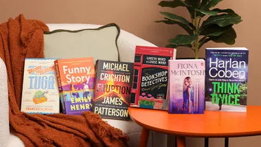A collection of six colorful books displayed on a chair and table with a plant in the background.