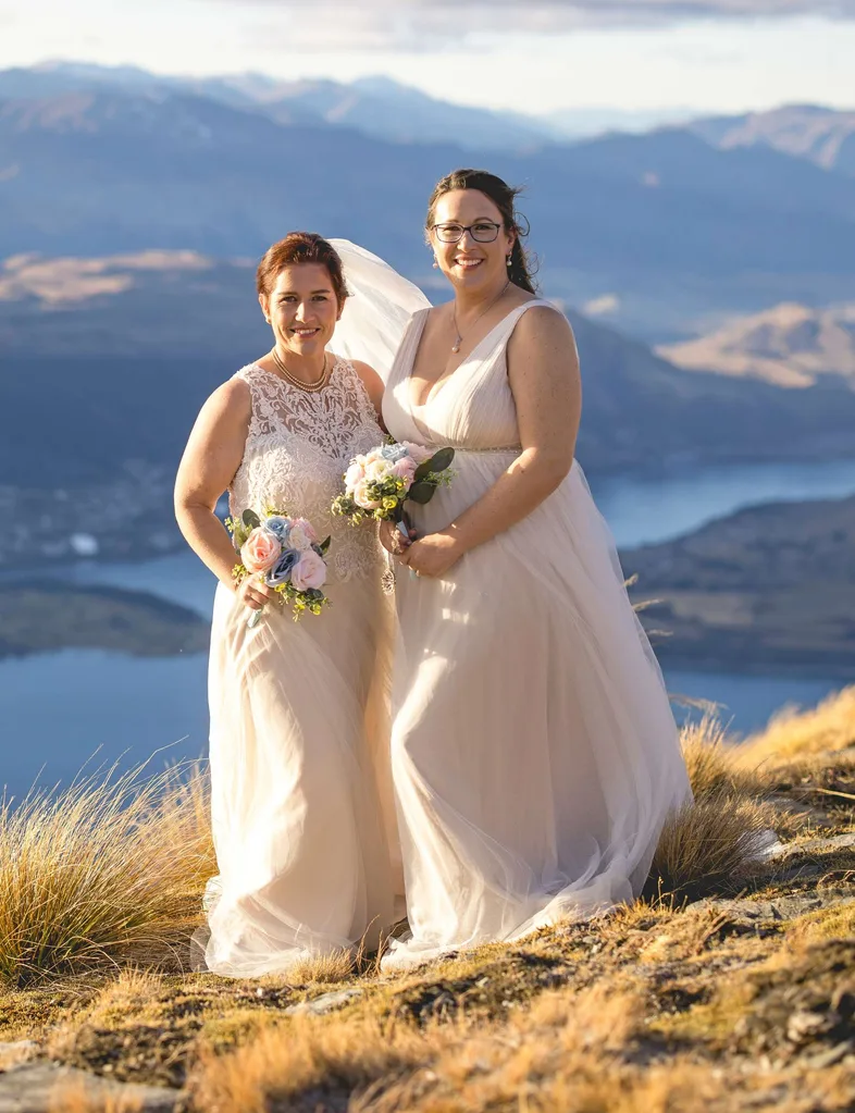 Melanie and Lead in their wedding dresses on the top of a mountain, a large body of water behind them