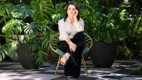 Melanie Lynskey sitting on a chair in front of tall plants, leaning her elbow on her knee