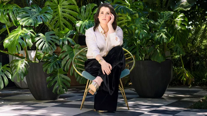 Melanie Lynskey sitting on a chair in front of tall plants, leaning her elbow on her knee