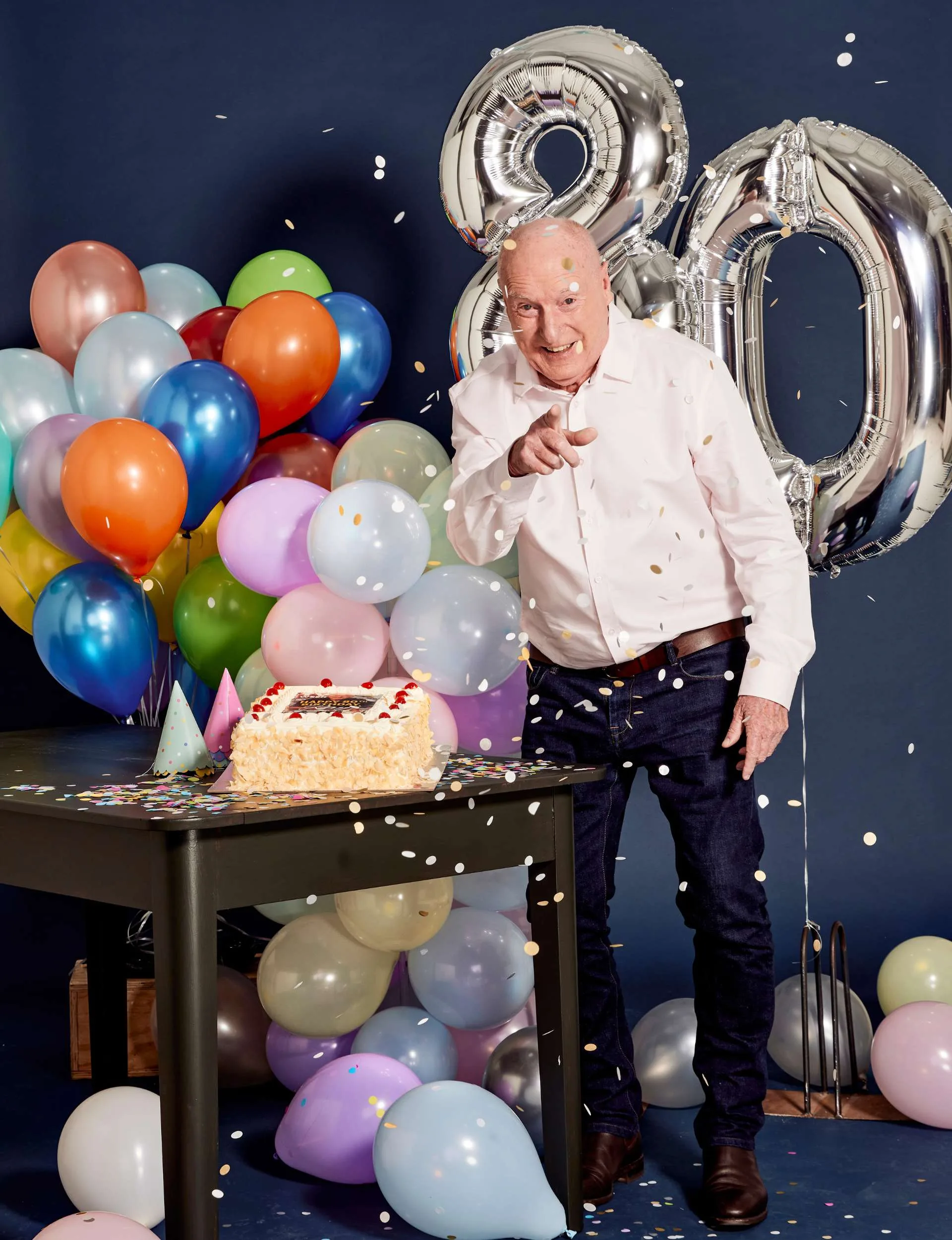 Ray pointing and smiling at the camera beside a cake and a bunch of balloons as confetti falls around him