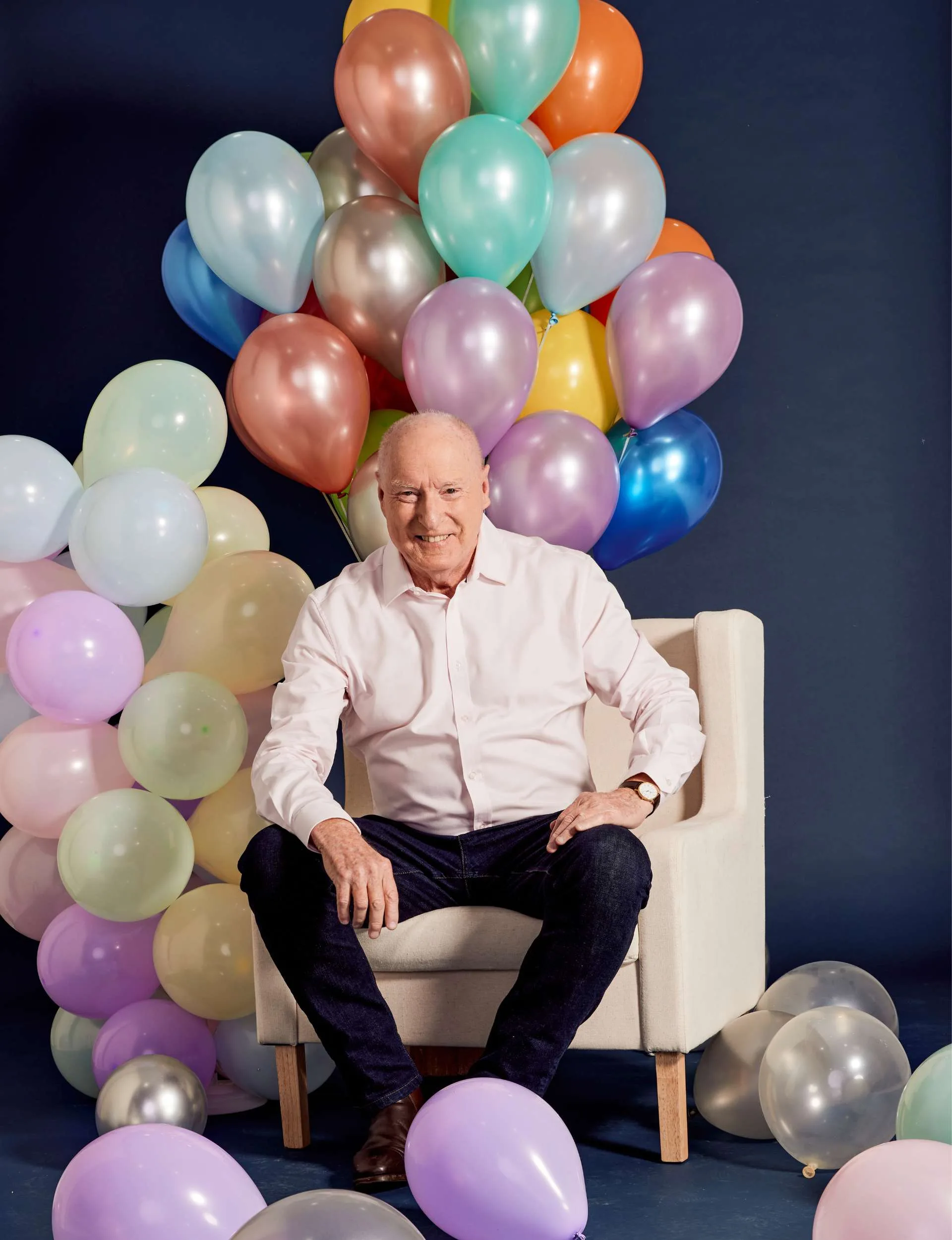Ray Meagher sitting on a chair in front of a bunch of colourful helium balloons
