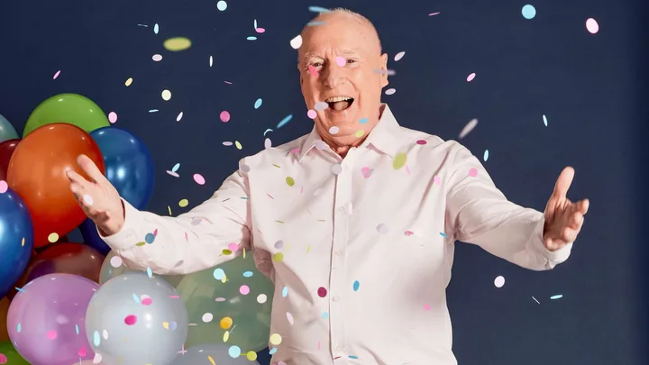 Ray Meagher standing in front of a bunch of balloons, throwing confetti in the air