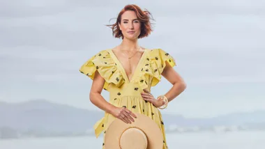 Samantha Hayes in a yellow dress holding a sunhat on a beach