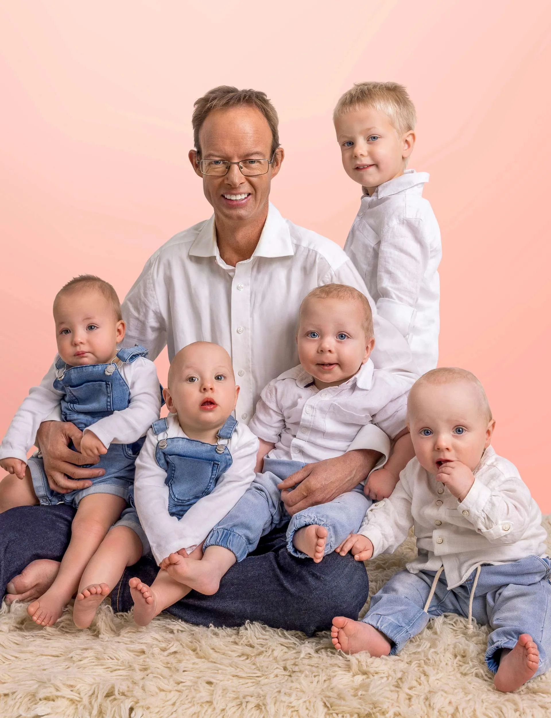 A family photo of Brett sitting on front of a light pink backdrop, with his five toddlers around him