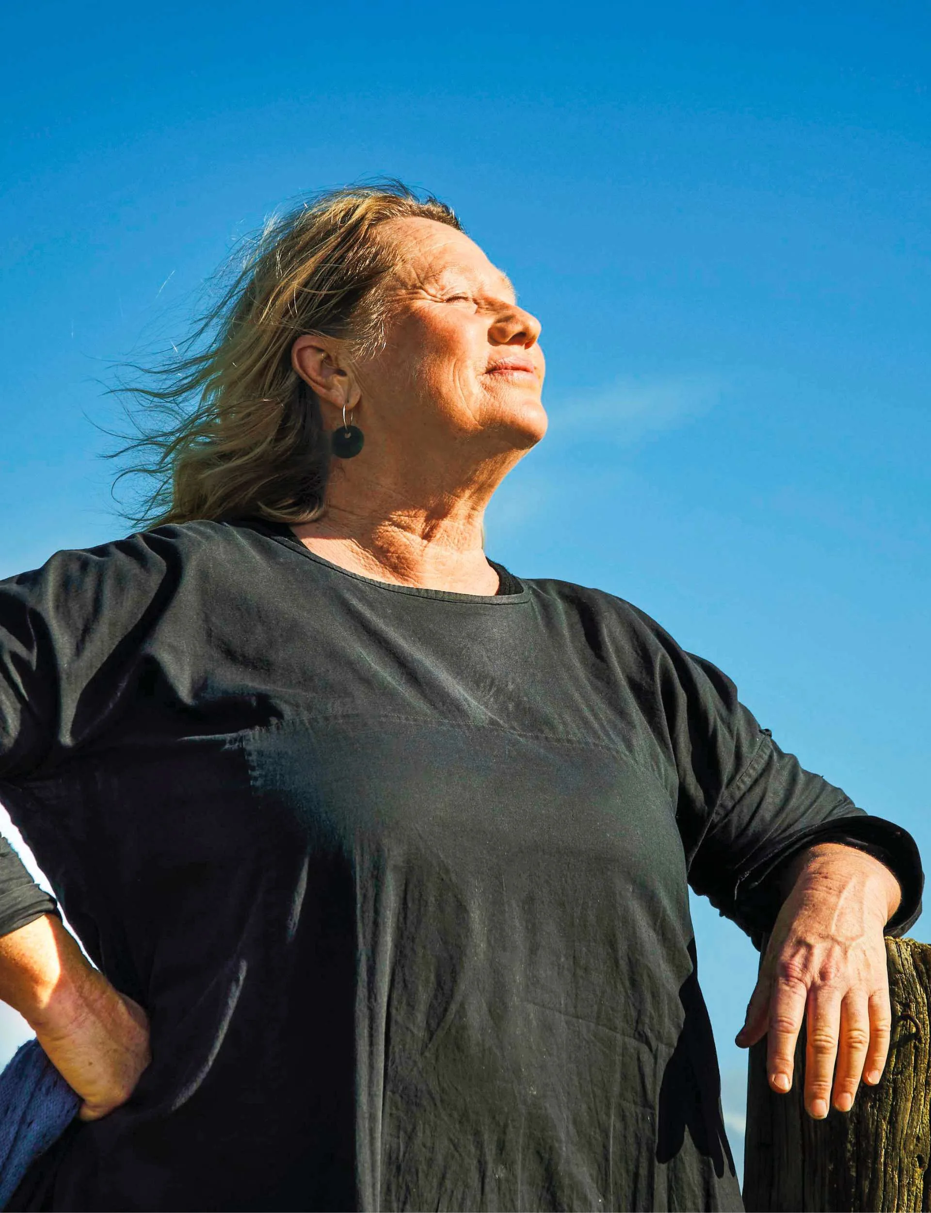 Nici Wickes leaning on a wooden post, head tilted up towards the clear sky, eyes closed and the wind blowing through her hair