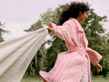 A person in a pink robe joyfully runs across a grassy area, trailing a white towel behind them.