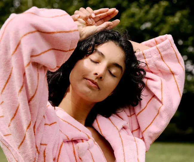 Woman in a pink striped robe with eyes closed, posing outdoors with hands above her head.
