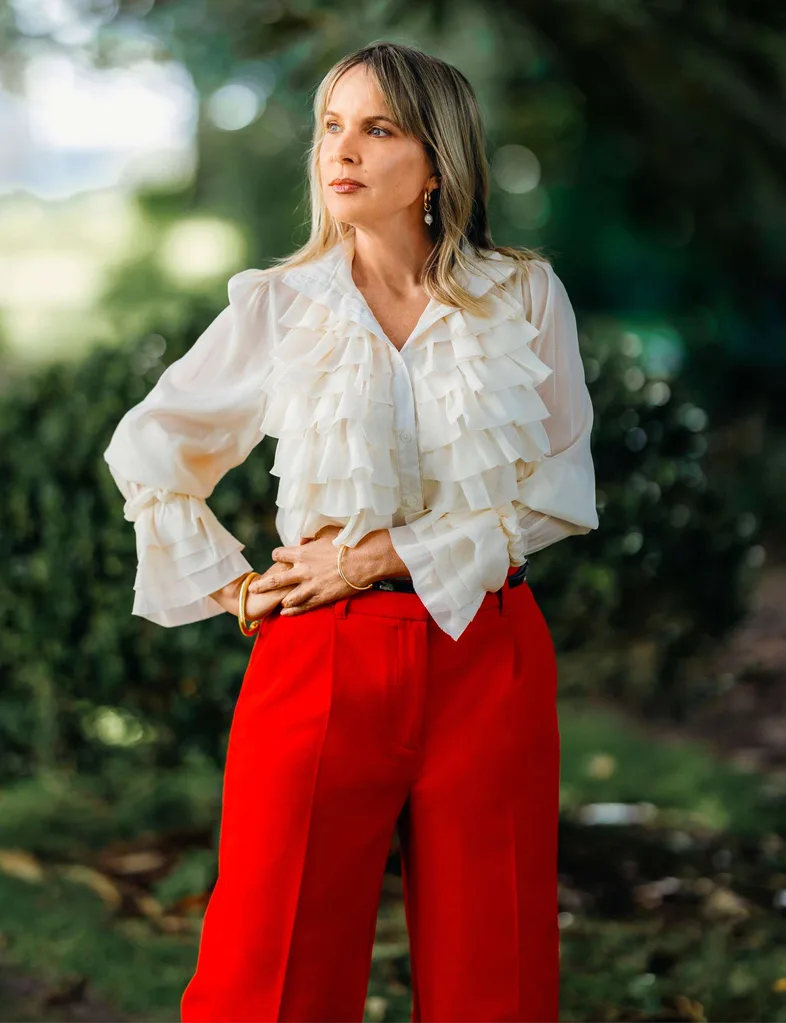 Angela in a white top and red pants posing in front of some trees