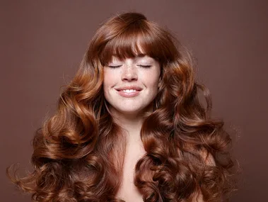 A smiling woman with long, curly, auburn hair and eyes closed, set against a brown background.