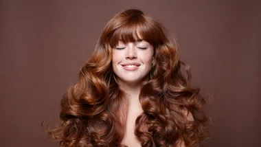 A smiling woman with long, curly, auburn hair and eyes closed, set against a brown background.