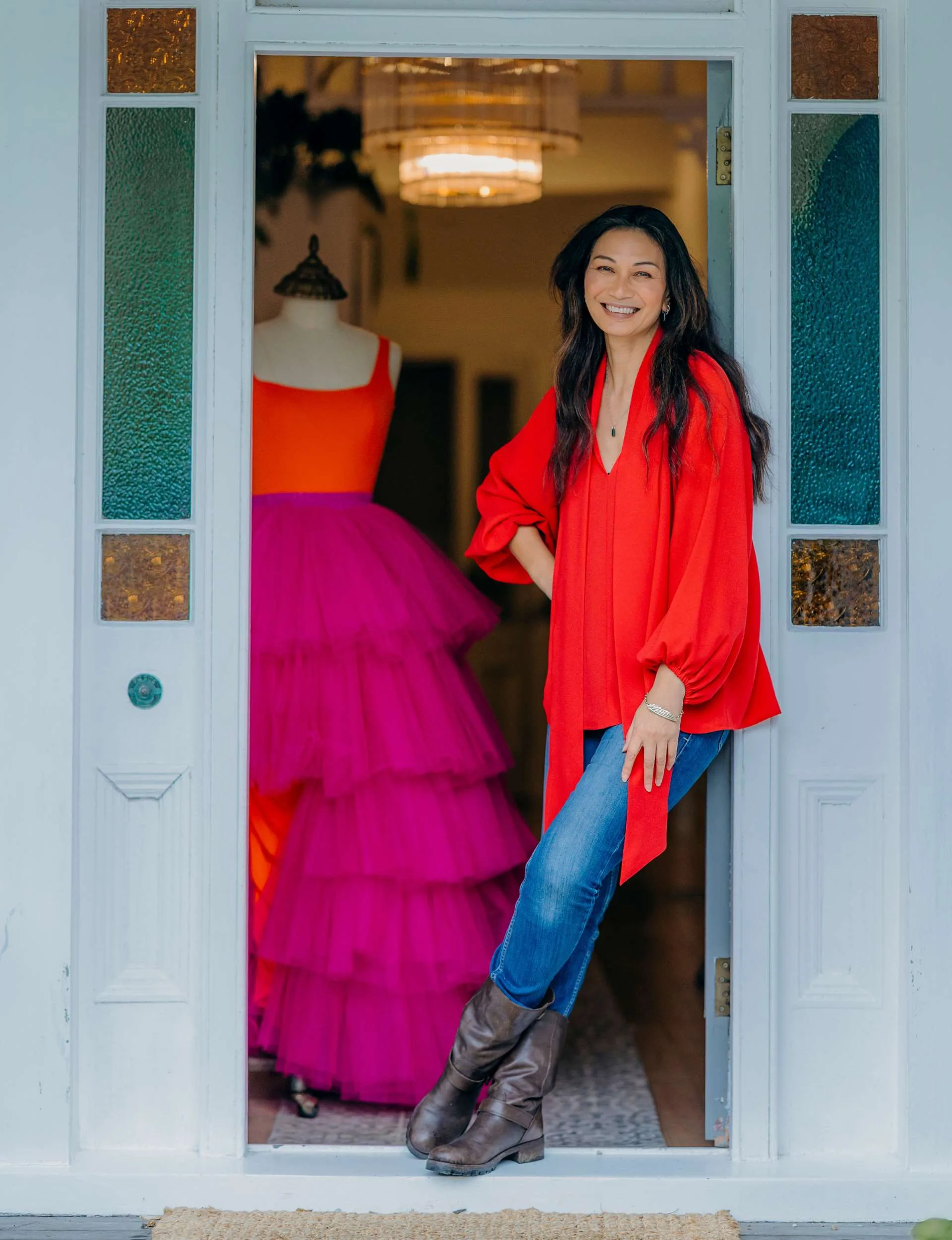 Boh Runga smiling from inside a doorway with a red and pink dress on a mannequin behind her