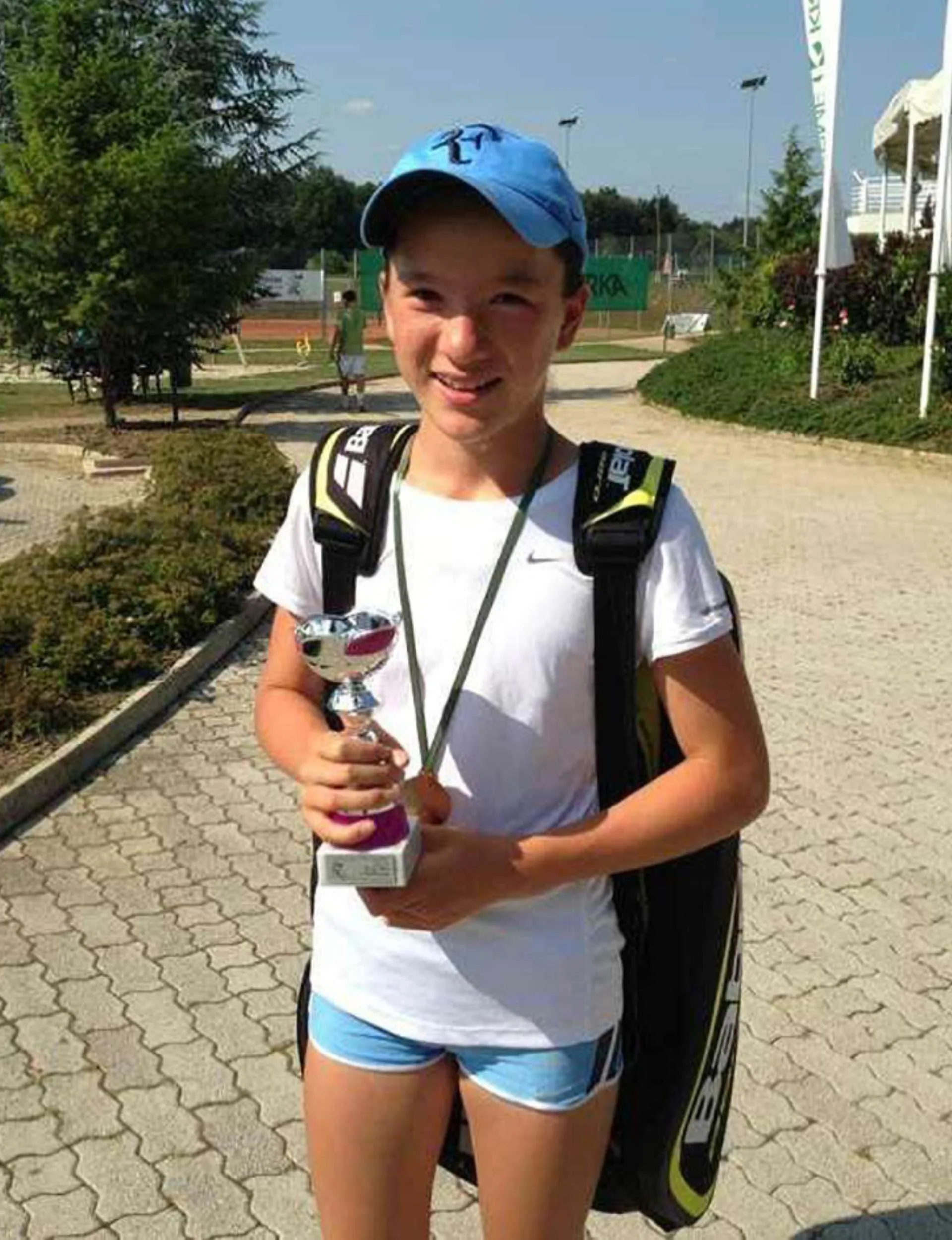 Lulu when she was young holding a small silver trophy, with a tennis bag on her back