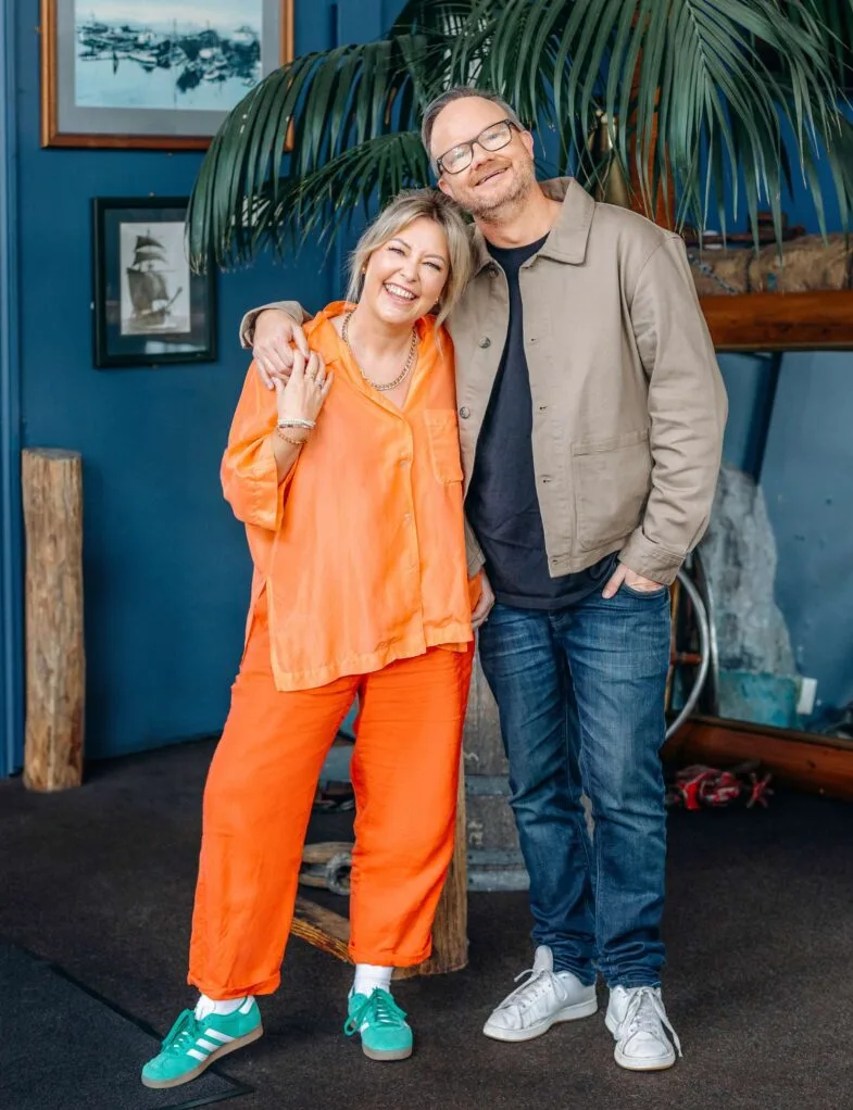 The pair posing in front of a large indoor plant