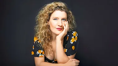 A woman with curly hair wearing a floral dress poses with her hand resting on her face against a dark background.