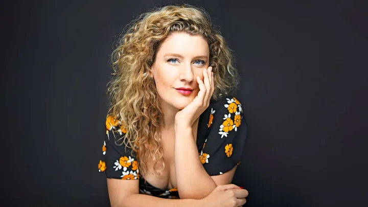 A woman with curly hair wearing a floral dress poses with her hand resting on her face against a dark background.