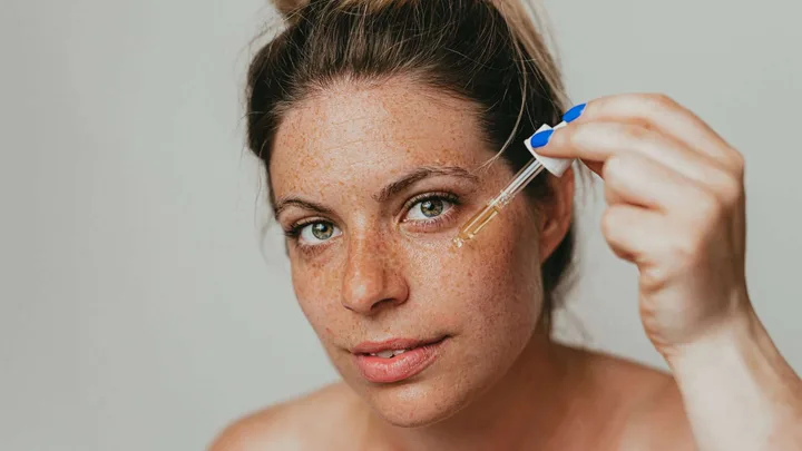 Woman applying facial serum with a dropper, close-up on her face against a neutral background.