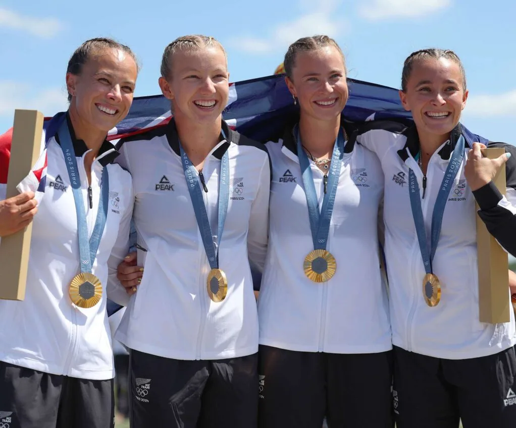 Olympic winners Lisa Carrington, Tara Vaughan, Olivia Brett and Alicia Hoskin wearing their medals, with an NZ flag over their shoulders