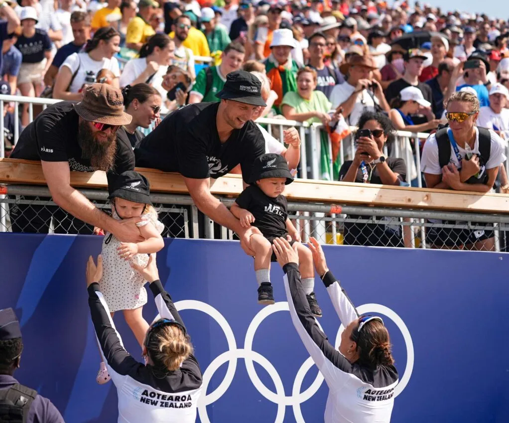 Golden women Brooke and Lucy reaching up to take their children from their dads up in the stands at the 2024 Olympic Games