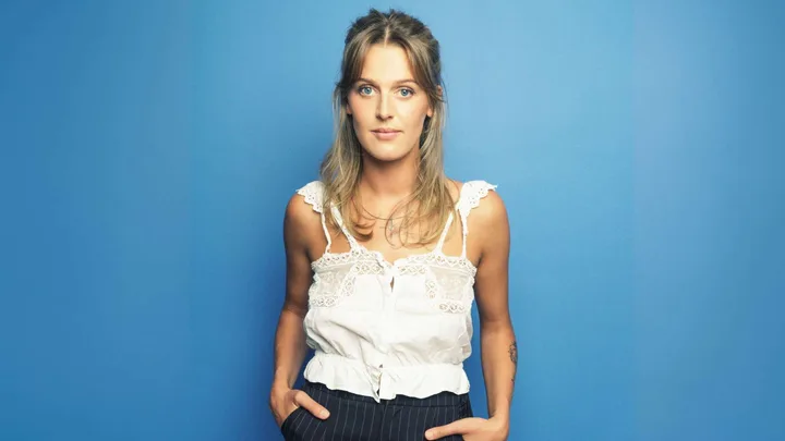 Moana McArtney with her hands in her pockets in front of a blue backdrop