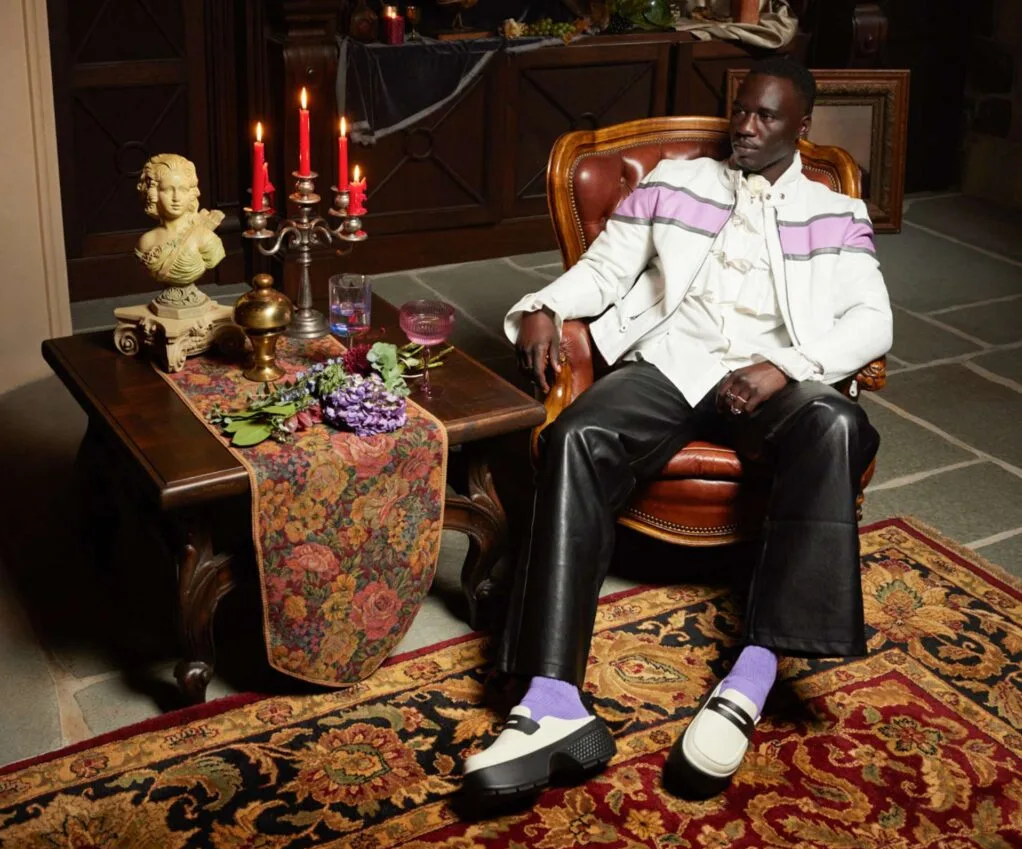 Male model wearing Crocs loafers with bright purple socks while sitting on an old-fashioned armchair