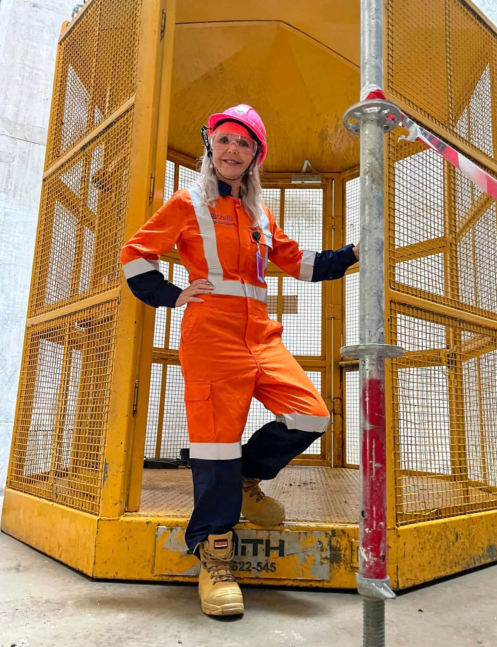 In a hi-vis jumpsuit, a pink hard hat and clear goggles on a construction site