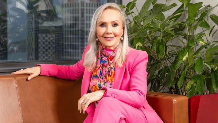 Desley Simpson sitting on a brown leather couch in a fluro pink suit