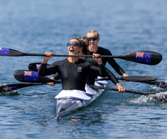 New Zealand women's kayak team celebrates winning, holding paddles with flags, wearing black uniforms on water.