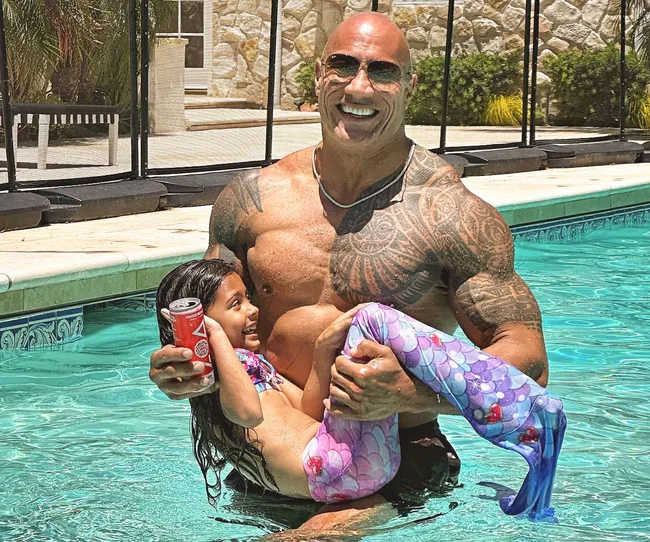 Man in sunglasses and swimwear holding a laughing child in a mermaid tail, standing in a pool with a drink can.