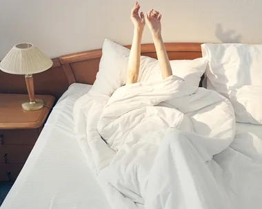 Person's arms stretch upward from white bed sheets on a bed with pillows and bedside lamps.