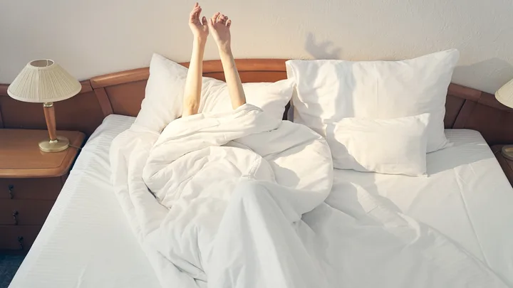 Person's arms stretch upward from white bed sheets on a bed with pillows and bedside lamps.