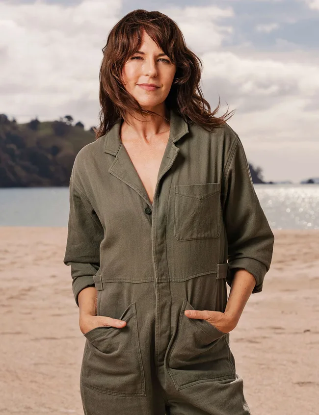 A person standing on a beach wearing an olive jumpsuit, with hands in pockets, against a cloudy sky and distant hills.