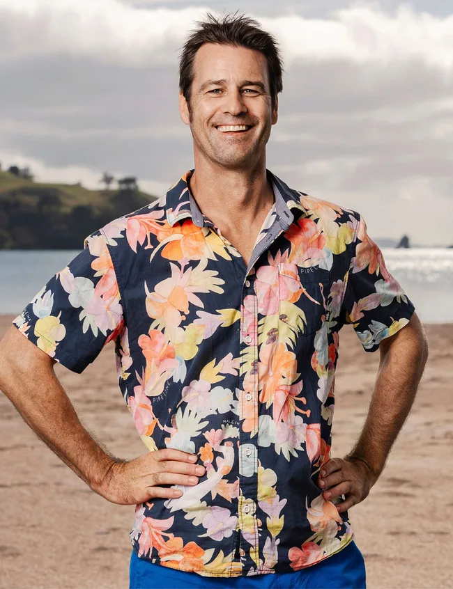 Man in a colorful floral shirt smiles on a beach with cloudy sky in the background.