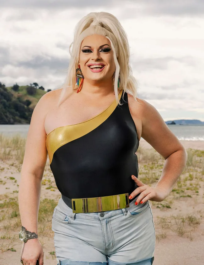 Blonde person in a one-shoulder black and gold top, smiling on a beach with cloudy sky in the background.