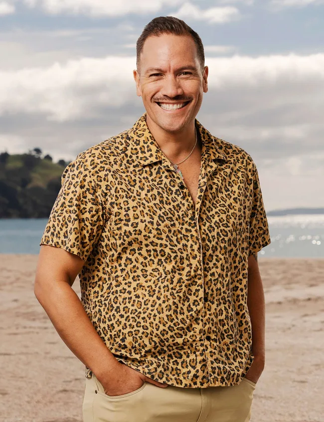 Man smiling at beach, wearing a leopard-print shirt and beige pants, with hands in pockets under a cloudy sky.