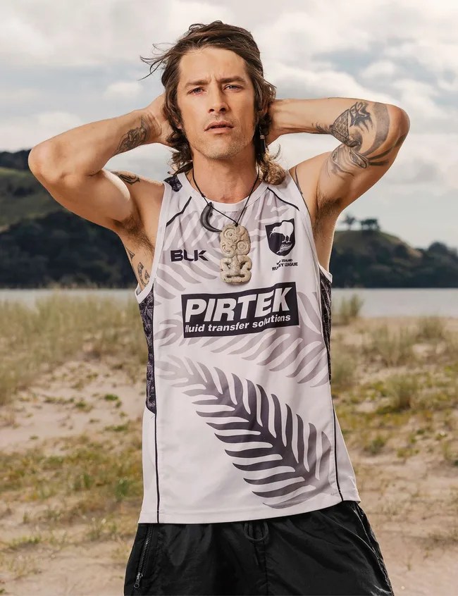 Man in a Pirtek sports tank top with tattoos on his arms stands outdoors, posing with hands behind head.