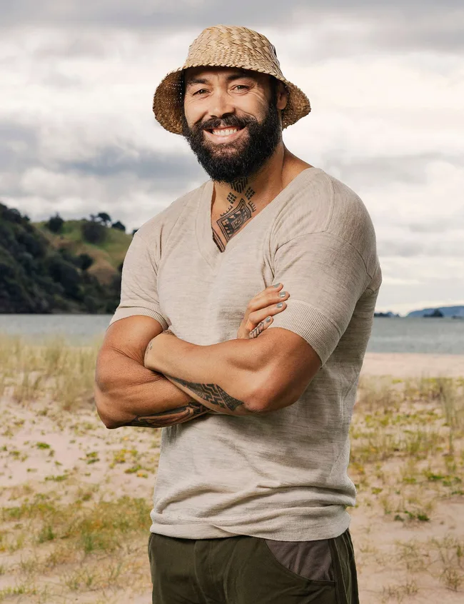 Man with beard and straw hat smiling with crossed arms on a beach.