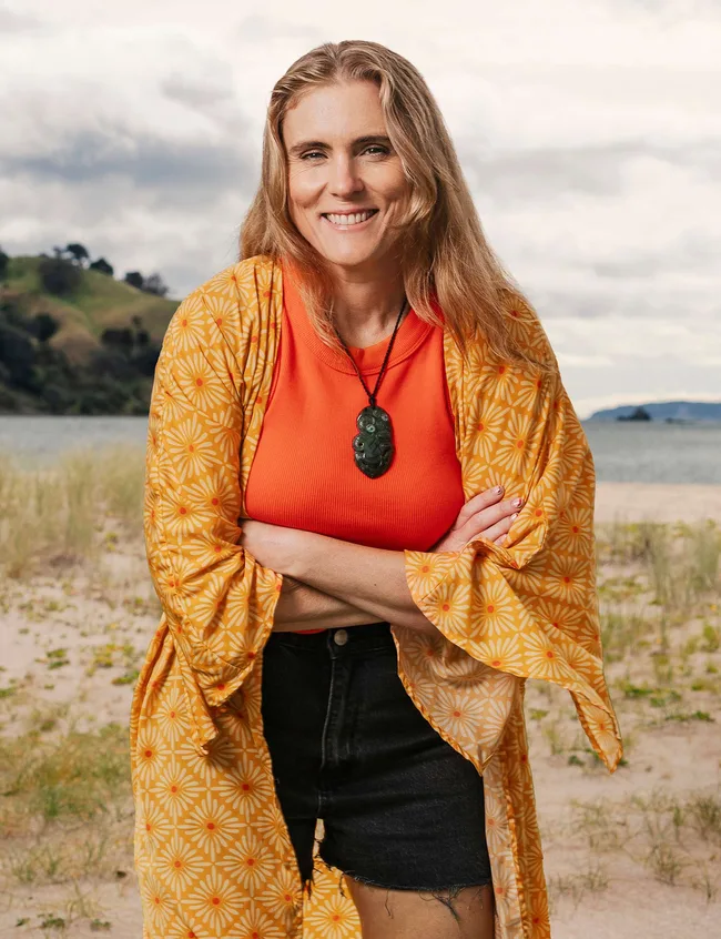 Woman in orange top and yellow floral wrap smiling, standing on a sandy beach with hills in the background.