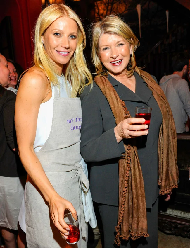 Two women smiling, holding drinks, one wearing a light apron, surrounded by people in a lively indoor setting.