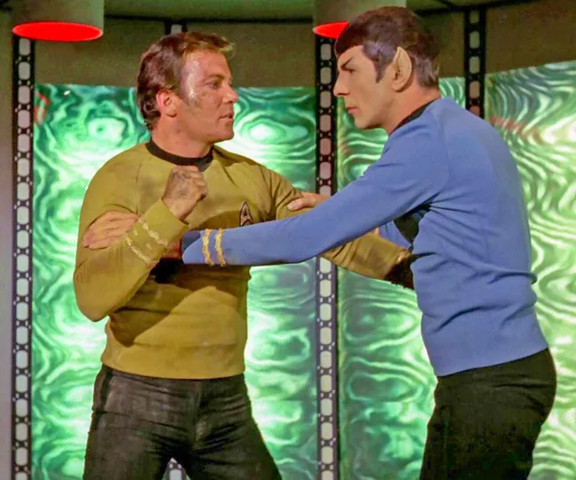 Two characters from Star Trek, one in yellow and one in blue, face each other in a tense moment on a spaceship.