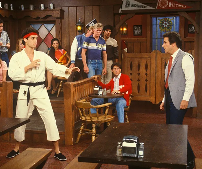 A man in a karate gi with a red headband faces another man in a restaurant, surrounded by onlookers.