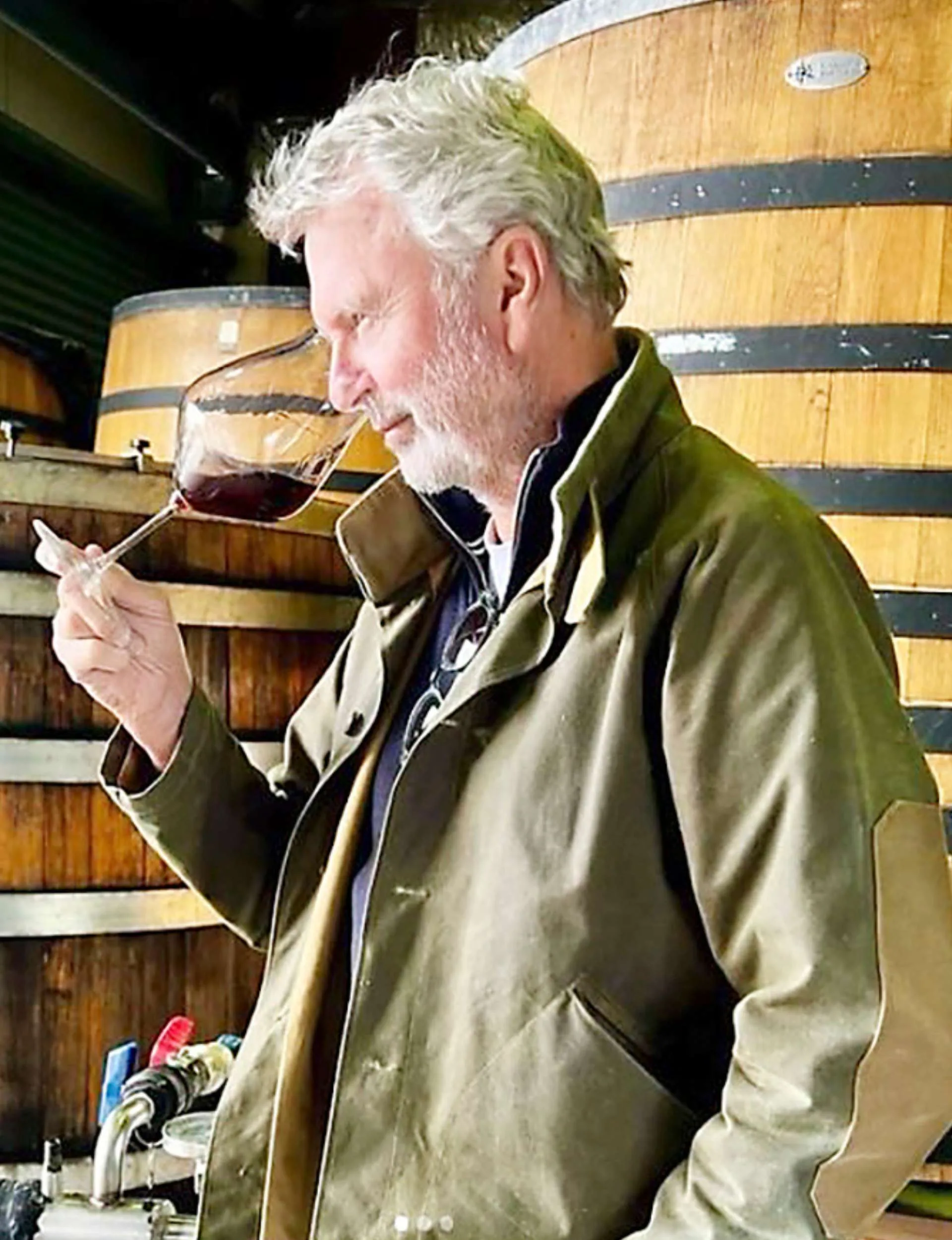 Sam Neill smelling a glass of wine in front of wine barrels