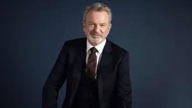Sam Neill in a suit in front of a dark backdrop