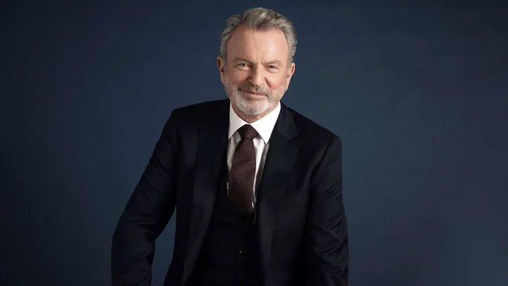 Sam Neill in a suit in front of a dark backdrop