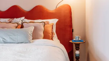 Cozy bedroom with orange headboard, decorative pillows, and a small side table holding a mug and book.