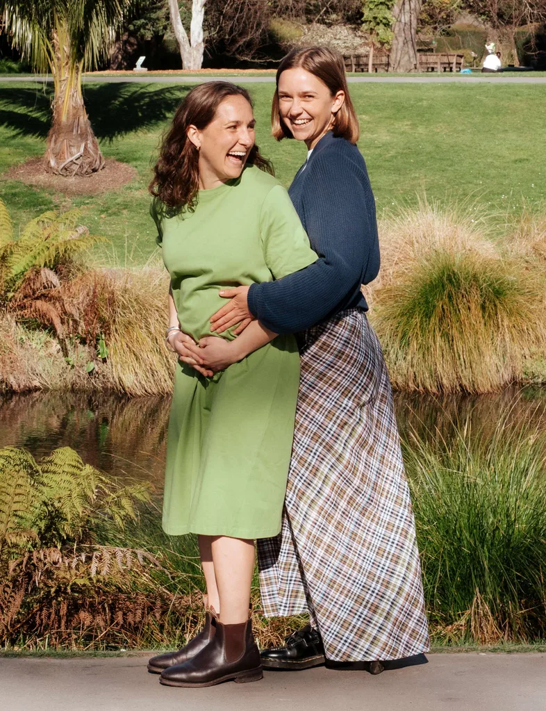 Two Raw Sisters Rosa and Margo in front of a river, Rosa cradling her baby bump and Margo reaching around to touch the belly too