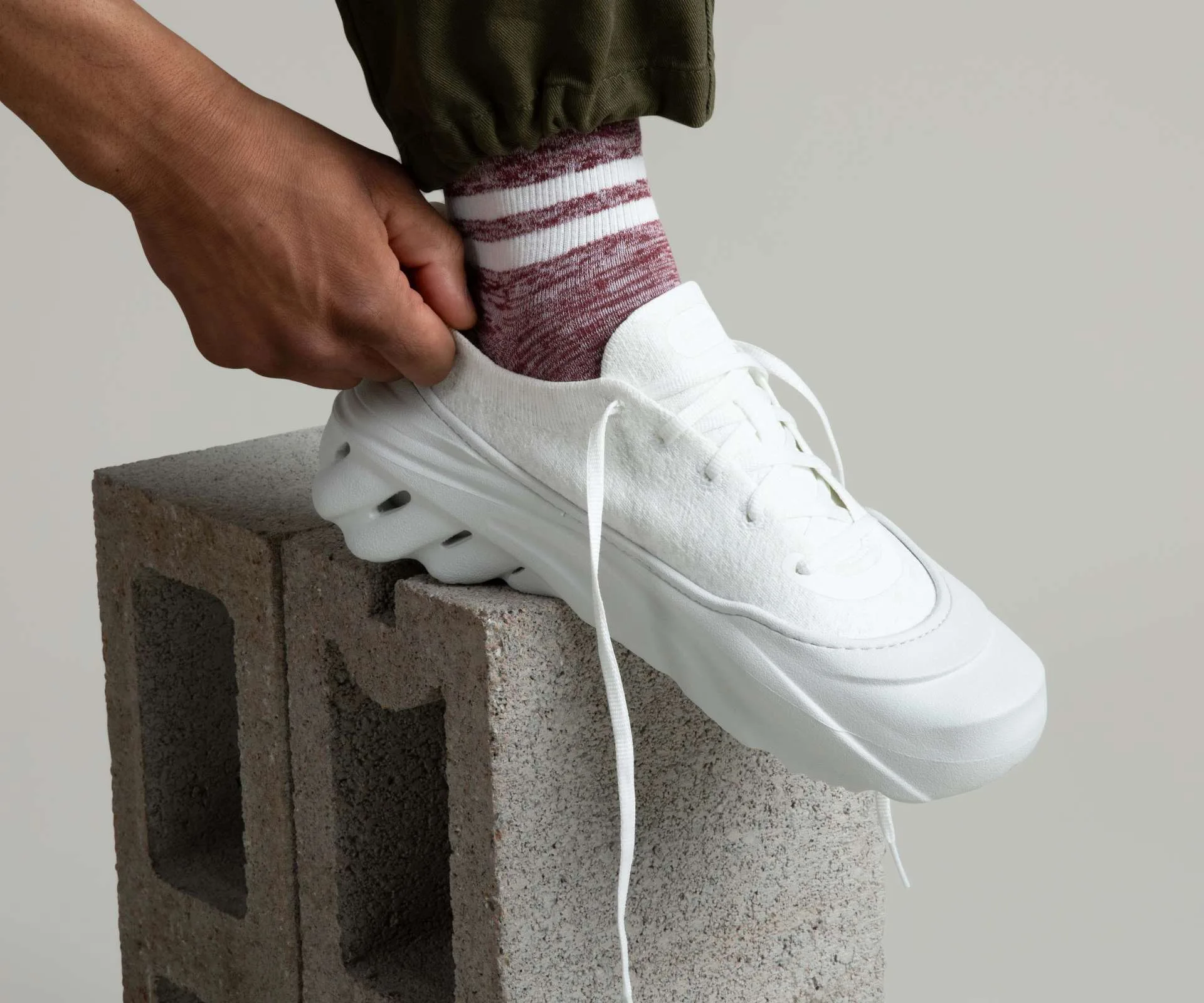 A male model pulling on a white crocs sneaker while propping his foot up on a cinderblock