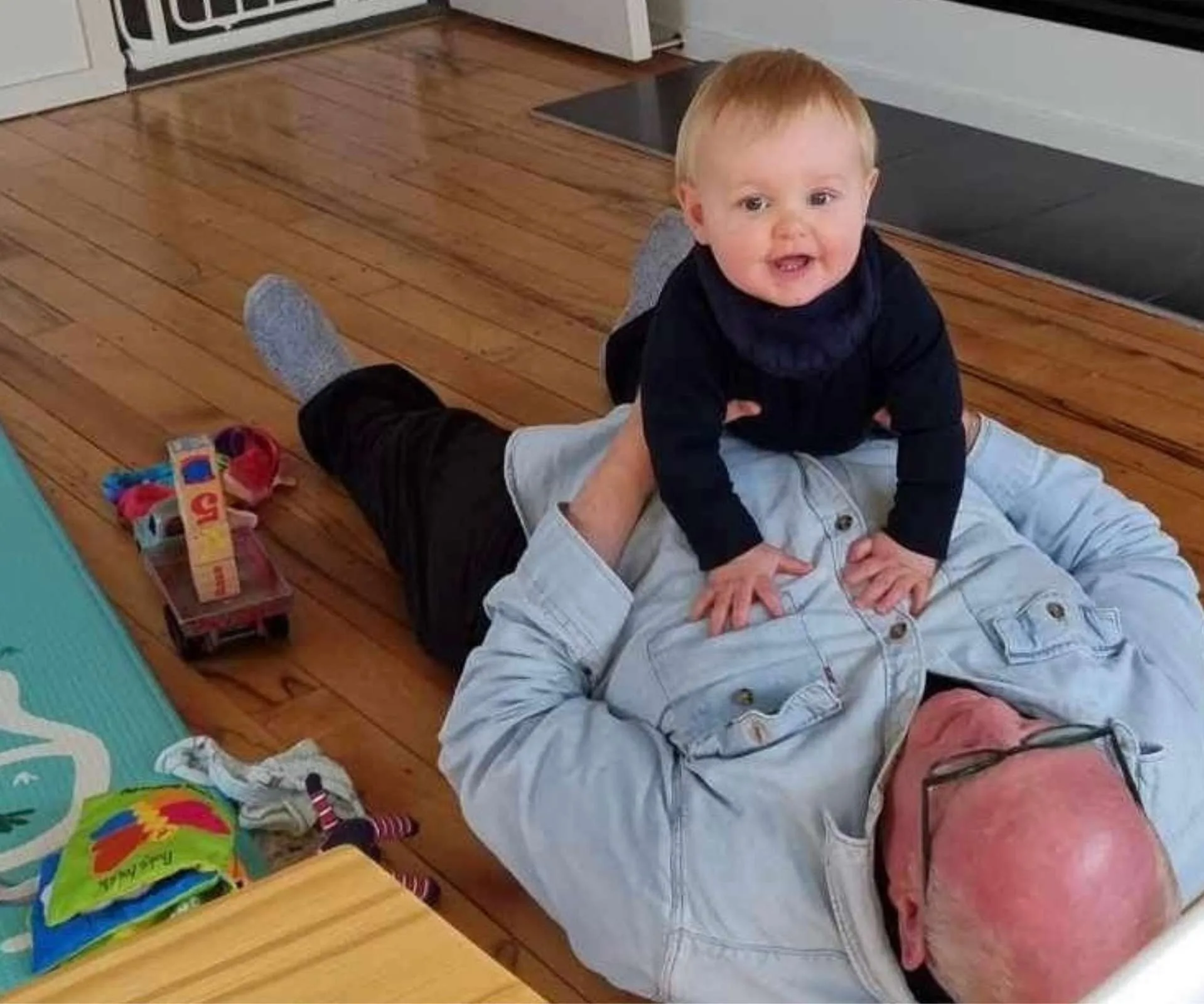 granddad Kevin Milne laying in his back while toddler Benny crawls over him