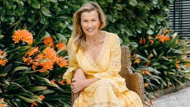 Robyn Malcolm wearing a yellow dress in front of some orange flowers